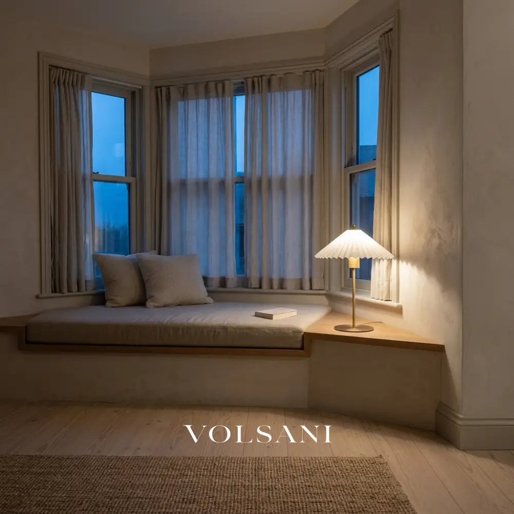 Brushed gold pleated-shade lamp lit on a bay window seat at dusk