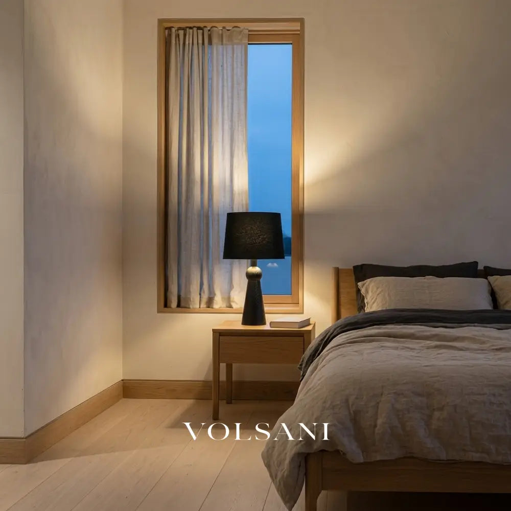 Black-shade Scandinavian wood table lamp lit on a bedside table in a Japandi bedroom at dusk
